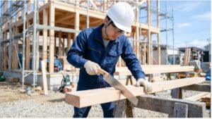 建築中の木造の骨組みの前で、ヘルメットと作業着の大工が木材をのこぎりで切っている