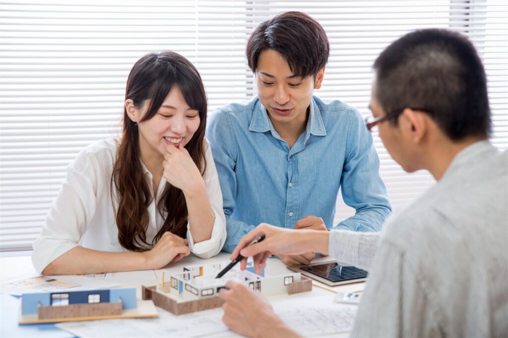 机の上の住宅の模型と図面を見ながら、夫婦と設計士が打ち合わせをしている