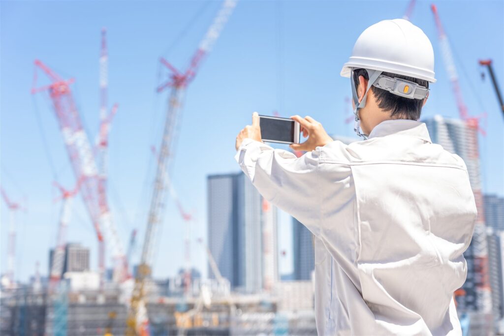 ヘルメットと作業服の人が後ろ向きにスマホで建設現場を撮る様子